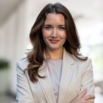 Professional headshot of a smiling woman with brown hair wearing a light beige blazer, neutral office background, arms crossed lightly.