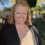 Smiling woman with curly blonde hair wearing a beige top and black cardigan outdoors in sunlight.