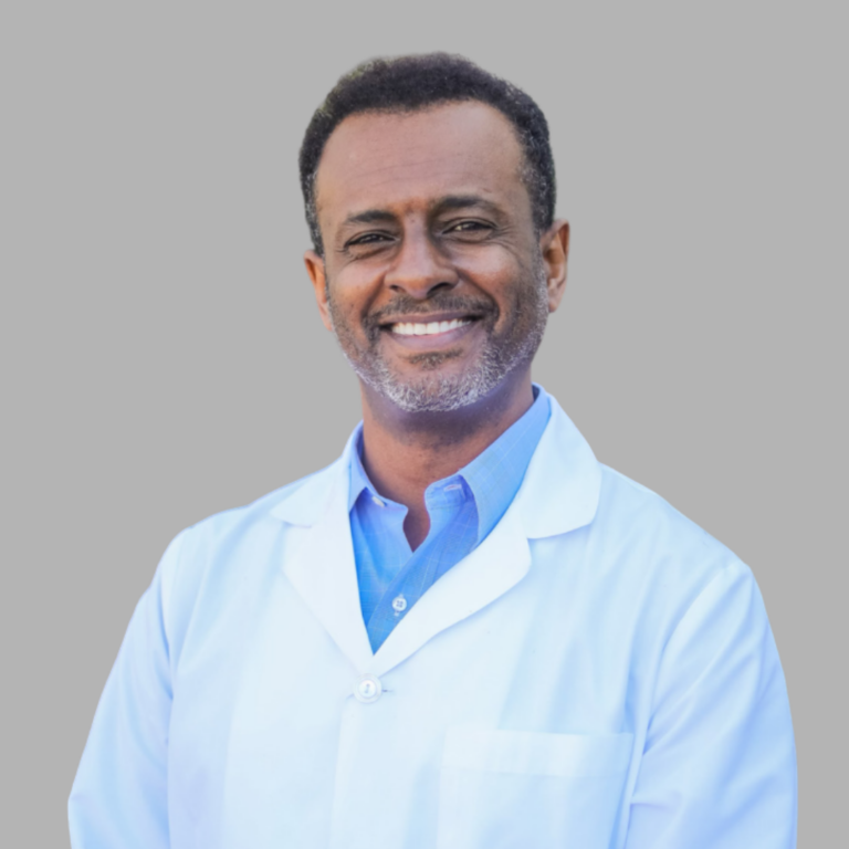 Smiling male doctor in a light blue lab coat and blue shirt, against a gray background.