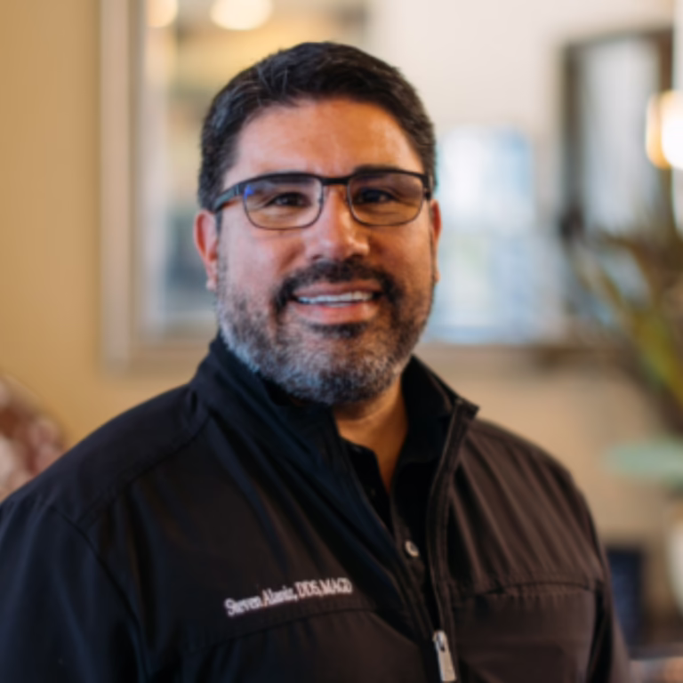 Smiling man with dark hair and glasses wearing a black zip-up jacket indoors.