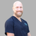 Smiling male healthcare professional wearing navy scrubs against a gray background.
