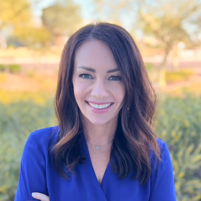Smiling woman with brown hair in a blue blouse, outdoors in a sunlit park background.