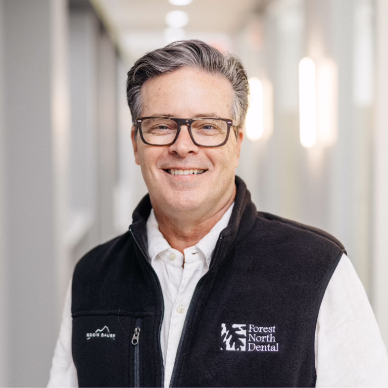Smiling middle-aged man with gray hair and glasses, wearing a black vest with the Forest North Dental logo, standing in a bright hallway.