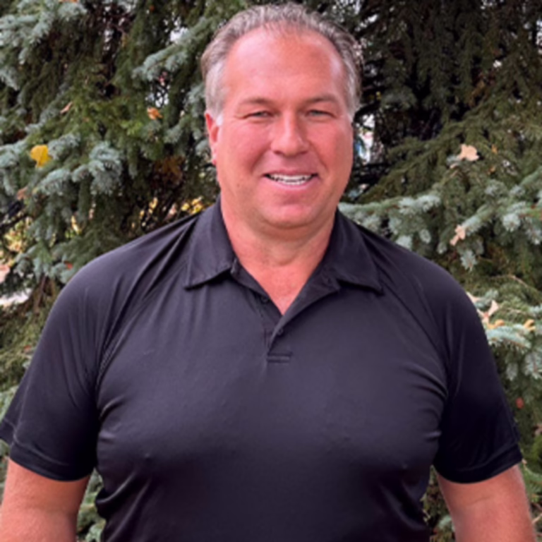 Smiling man in a black polo shirt standing outdoors with evergreen trees in the background.