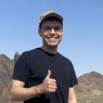 Smiling man wearing a tan cap and black t-shirt gives a thumbs up outdoors with rocky mountains in the background.