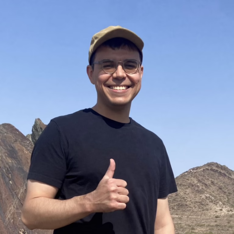 Smiling man wearing a tan cap and black t-shirt gives a thumbs up outdoors with rocky mountains in the background.