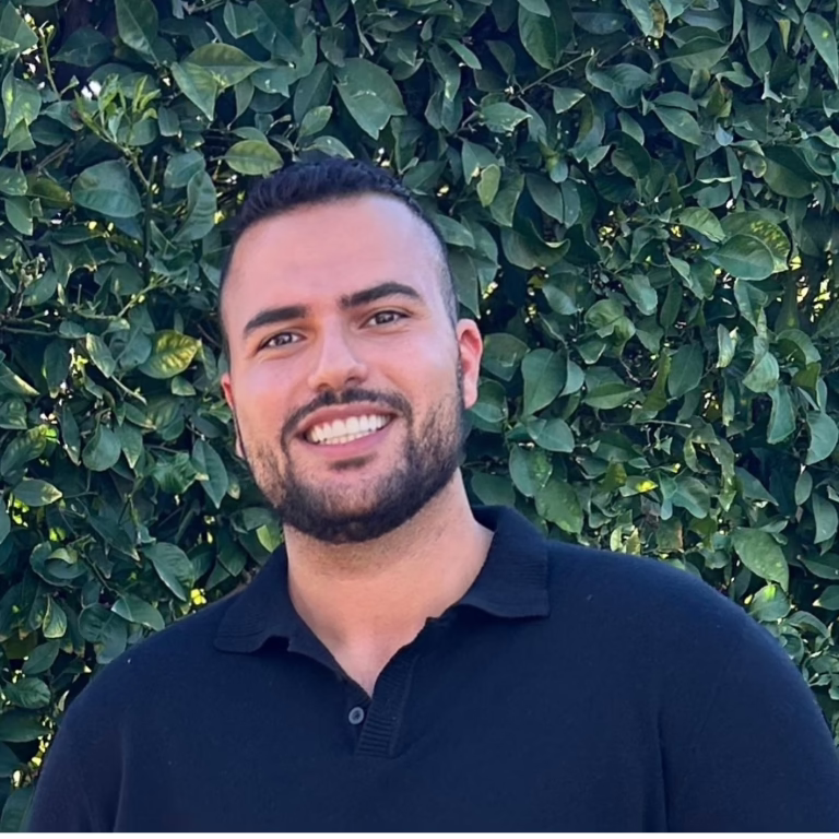 Smiling man with short dark hair and beard in a navy polo, standing in front of a green leafy hedge.