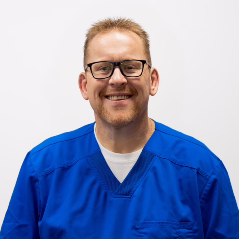Smiling healthcare professional wearing blue scrubs and glasses against a white background.