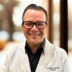 Smiling man wearing black-framed glasses and a white dental coat with 'Dr. Michael Augustine Dentistry' embroidered on the chest.