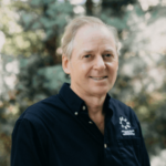 Smiling older man with light hair wearing a navy button-down shirt, standing outdoors with a left-chest logo visible.