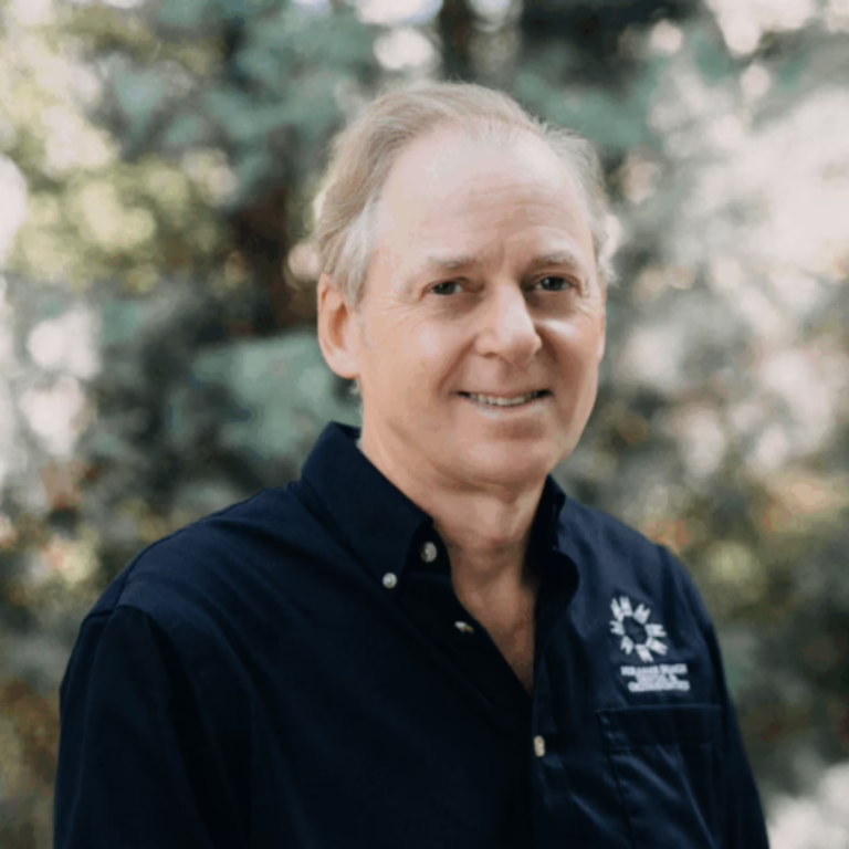 Smiling older man with light hair wearing a navy button-down shirt, standing outdoors with a left-chest logo visible.