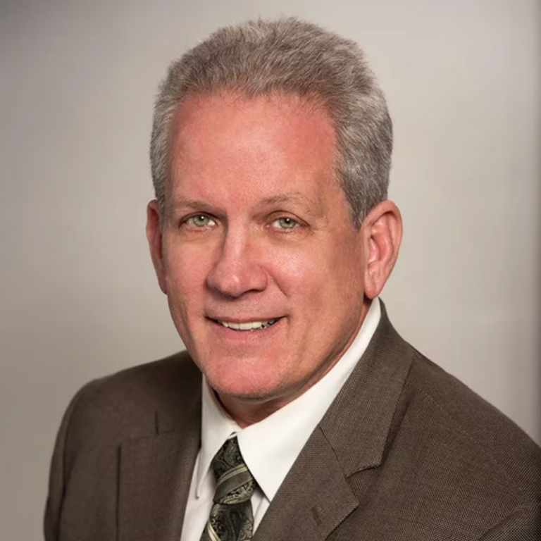 Head-and-shoulders portrait of a smiling man in a brown suit with a patterned tie, against a neutral beige background.