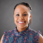 Portrait of a smiling woman with braided hair wearing a colorful patterned blouse against a gray background.