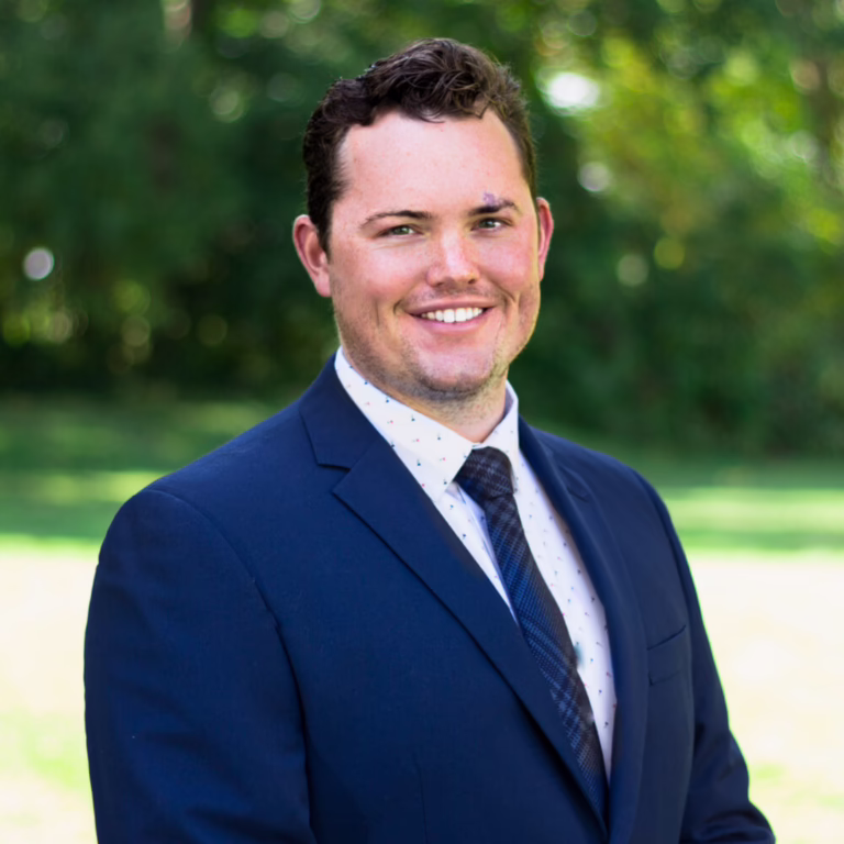 Professional headshot: smiling man in a navy suit outdoors with a green, blurred background.
