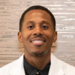 Smiling man in a white lab coat against a light wooden background.