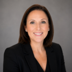 Professional headshot of a smiling woman in a black blazer against a neutral gray backdrop, businesslike and approachable.