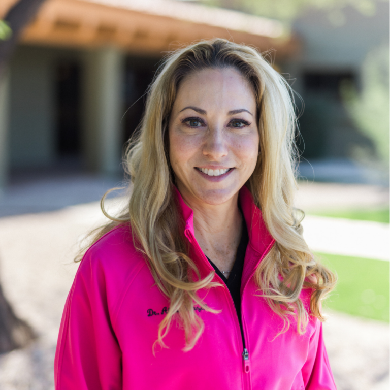 Smiling woman with long blonde hair wearing a bright pink jacket outdoors.