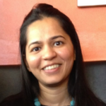 Smiling woman with long dark hair and turquoise bead necklace, photographed indoors against a coral wall.