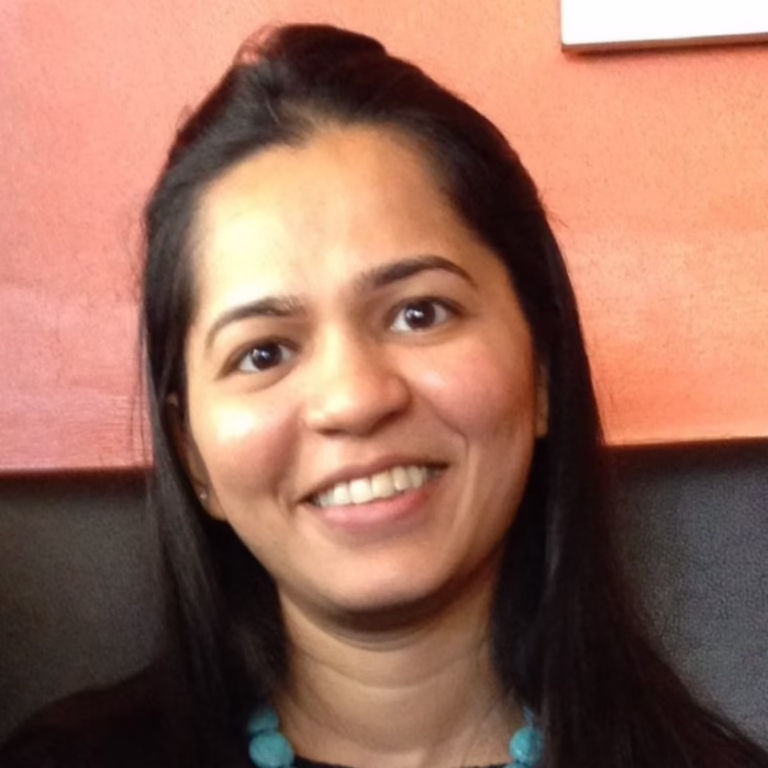 Smiling woman with long dark hair and turquoise bead necklace, photographed indoors against a coral wall.