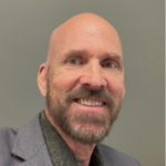 Smiling bald man with gray beard wearing a gray blazer, looking at the camera as a close-up portrait.
