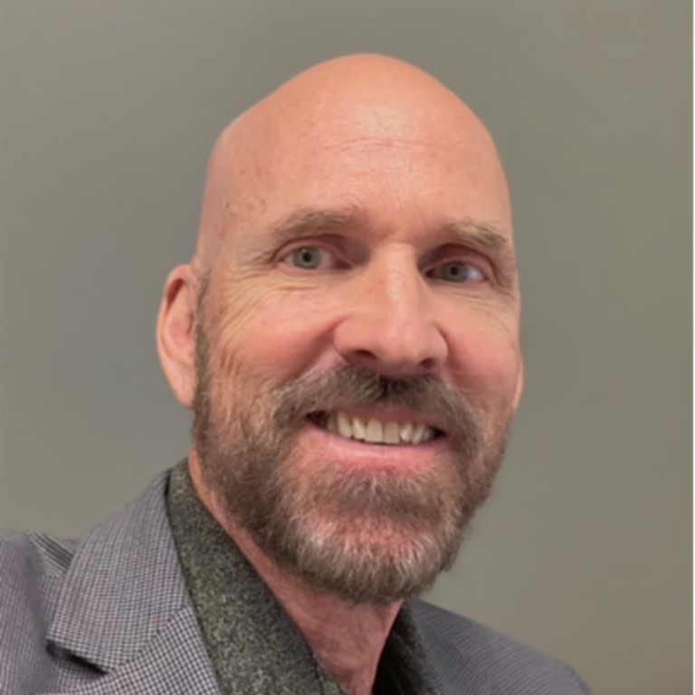 Smiling bald man with gray beard wearing a gray blazer, looking at the camera as a close-up portrait.