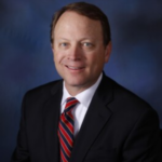 Professional headshot of a man in a dark suit and red striped tie, smiling against a blue backdrop.