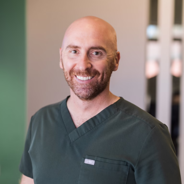 Smiling bald man in dark green scrubs posing in a clinic hallway, looking at the camera: portrait of a healthcare professional