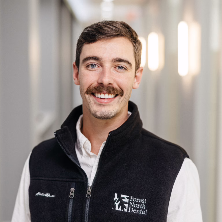 Smiling man in a white shirt and black zip vest with Forest North Dental logo, portrait in a bright hallway.