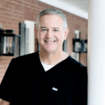 Smiling man in black scrubs standing near a white pillar in a brick-walled clinic.