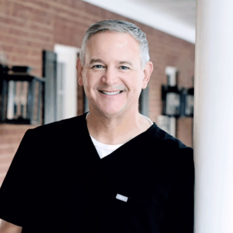 Smiling man in black scrubs standing near a white pillar in a brick-walled clinic.