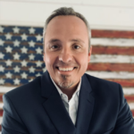 Professional headshot of a smiling man in a navy suit, with an American flag backdrop behind him.