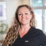 Smiling woman with long wavy hair in black scrubs, standing in a bright clinic setting