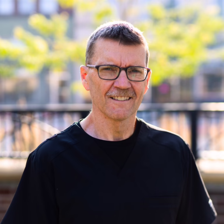 Portrait of a middle-aged man with glasses smiling, outdoors, wearing black scrubs.