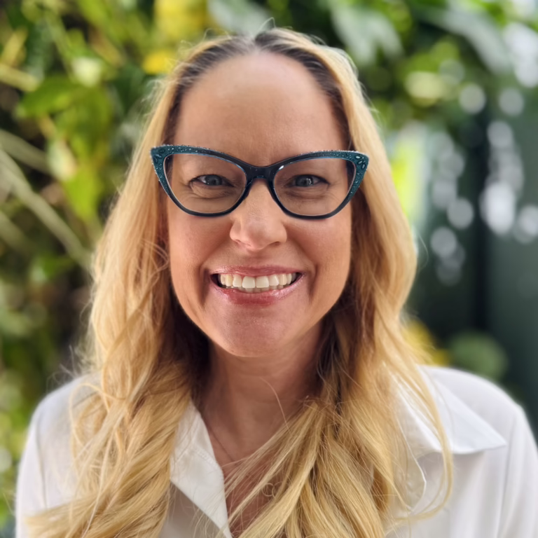 Smiling woman with blonde hair and cat-eye glasses outdoors, wearing a white shirt.