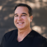 Smiling man with short dark hair wearing a black V-neck shirt, outdoors with a blurred background.