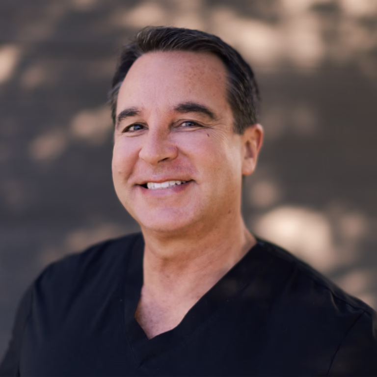 Smiling man with short dark hair wearing a black V-neck shirt, outdoors with a blurred background.