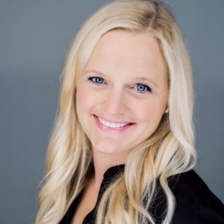 Head-and-shoulders portrait of a smiling blonde woman with blue eyes wearing a black top against a gray background.