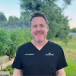 Smiling man in black scrubs standing in a garden with green plants behind him.