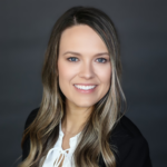 Head-and-shoulders portrait of a smiling professional woman with blue eyes and long wavy light-brown hair, wearing a black blazer and white blouse against a dark gray background.