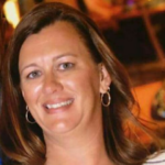 Smiling woman with light brown hair, wearing hoop earrings, in warm indoor lighting.