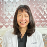 Portrait of a smiling female dentist in a white coat, with 'Chu DDS' embroidered on it, standing in front of a pink, patterned background.
