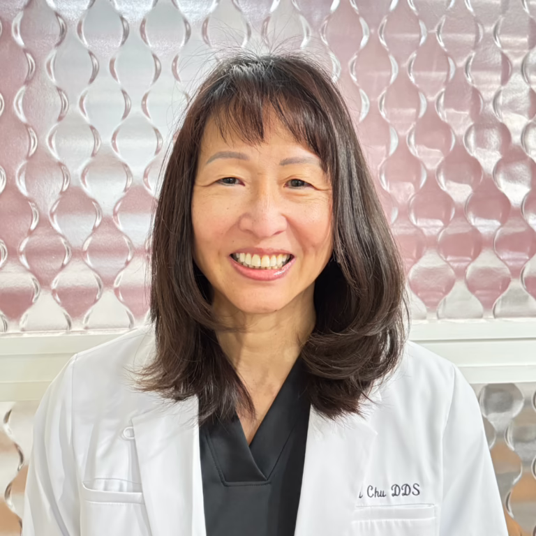 Portrait of a smiling female dentist in a white coat, with 'Chu DDS' embroidered on it, standing in front of a pink, patterned background.