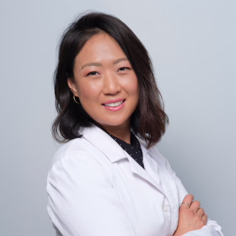 Portrait of a smiling Asian woman in a white lab coat with arms crossed against a light gray backdrop, conveying professionalism and approachability.