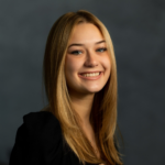 Smiling young woman with long blonde hair in a black top against a dark backdrop, portrait-style headshot for a professional profile.
