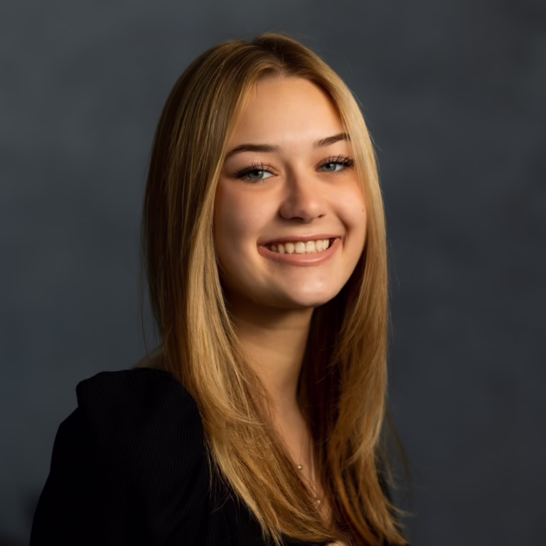 Smiling young woman with long blonde hair in a black top against a dark backdrop, portrait-style headshot for a professional profile.