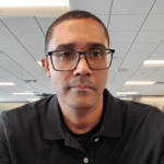 Portrait of a man wearing black glasses and a black polo shirt in an office setting, looking at the camera.