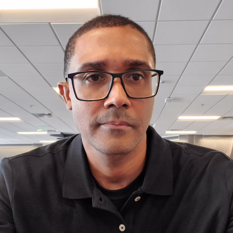 Portrait of a man wearing black glasses and a black polo shirt in an office setting, looking at the camera.