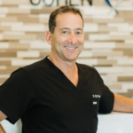 Smiling male healthcare professional in black scrubs leaning on a white counter with a light tile wall background