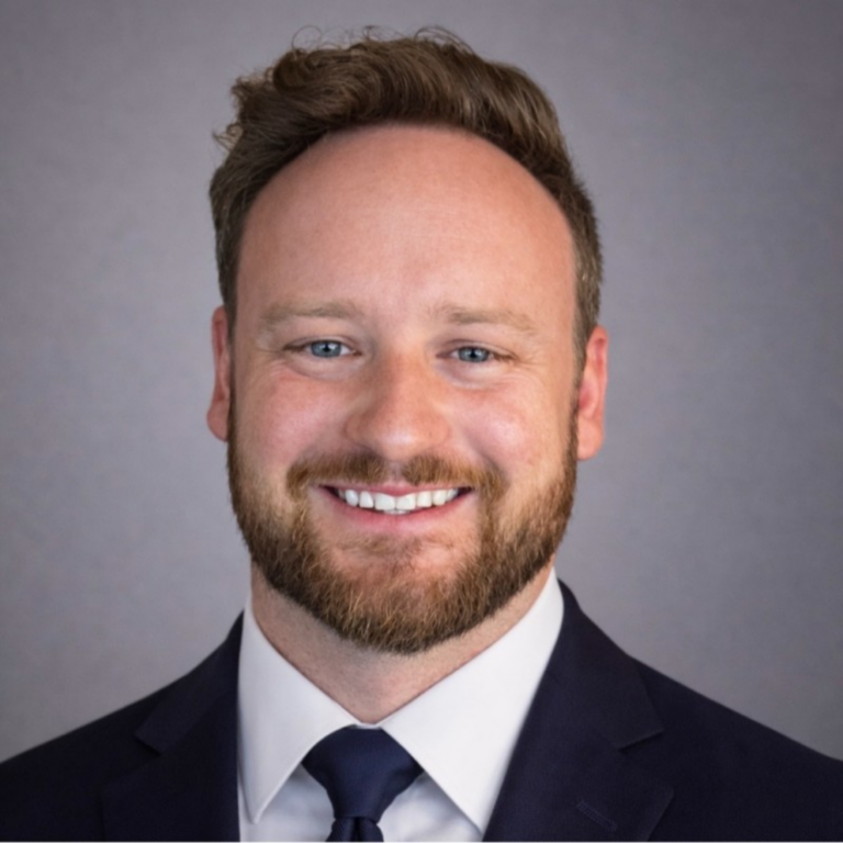 Smiling man with light brown hair and beard, wearing a navy suit and white shirt against a gray backdrop.