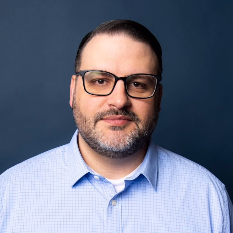 Portrait of a man with dark hair, glasses, and a short beard, wearing a light blue checkered shirt against a dark blue background.